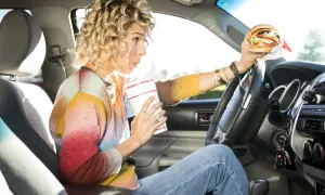 Frau trinkt und hält einen Burger beim Autofahren. Ob man beim Autofahren essen darf, ist eine Frage der Konzentration.