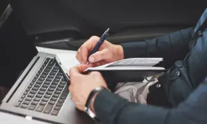 Mann schreibt im Notizbuch, während er im Auto sitzt. Er dokumentiert seinen Arbeitsweg, ob privat oder dienstlich, und nutzt dabei seinen Laptop.