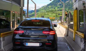 Schwarzer Firmenwagen an einer Mautstation, bereit für die Fahrt ins Ausland. Berge im Hintergrund.