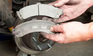 Hände halten einen neuen und einen abgenutzten Bremsbelag. Der alte Bremsbelag ist fast aufgebraucht, zeigt Verschleißspuren und ist mit Bremsstaub bedeckt.