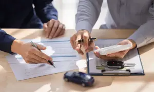 Zwei Personen besprechen Dokumente und eine Rechenmaschine. Ein Autoschlüssel und ein Spielzeugauto liegen auf dem Tisch. Sie klären den Listenpreis netto oder brutto.