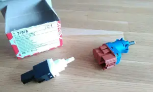 Zwei neue Bremslichtschalter liegen neben einer geöffneten Verpackung. Ein Teil ist blau und braun, das andere schwarz und weiß.