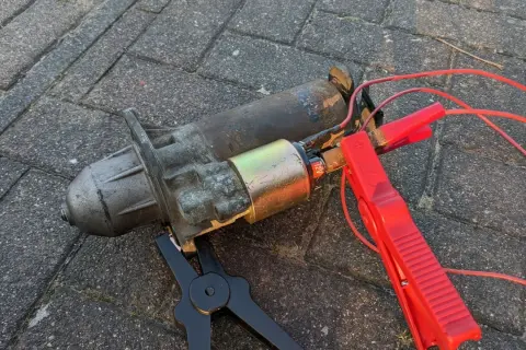 Ein defekter Anlasser, der nur klackt, liegt auf Pflastersteinen. Rote Kabel mit Klemmen sind angeschlossen.