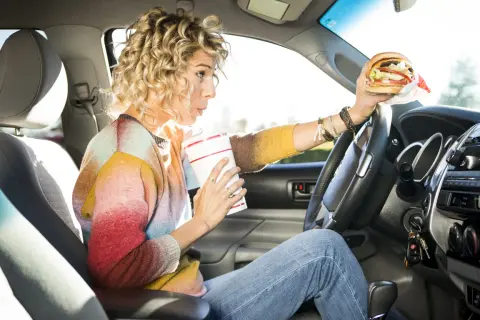 Frau trinkt und hält einen Burger beim Autofahren. Ob man beim Autofahren essen darf, ist eine Frage der Konzentration.