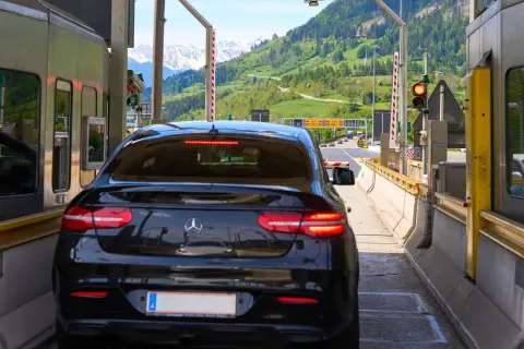 Schwarzer Firmenwagen an einer Mautstation, bereit für die Fahrt ins Ausland. Berge im Hintergrund.