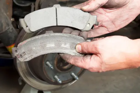 Hände halten einen neuen und einen abgenutzten Bremsbelag. Der alte Bremsbelag ist fast aufgebraucht, zeigt Verschleißspuren und ist mit Bremsstaub bedeckt.