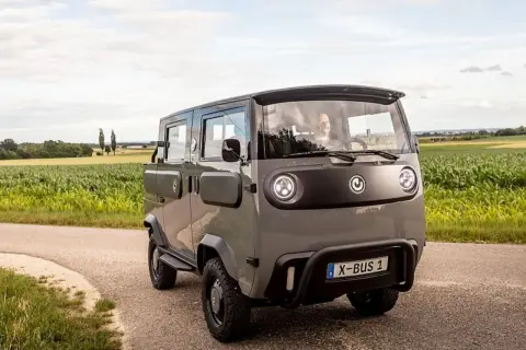 Grauer X-Bus 1 fährt auf Feldweg, Fahrer lächelt. Dieses Elektrofahrzeug ist ein Beispiel für moderne elektrische Verbraucher.