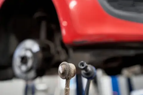 Zwei Koppelstangen liegen vor einem roten Auto. Die Koppelstange ist ein wichtiges Bauteil für die Fahrstabilität.