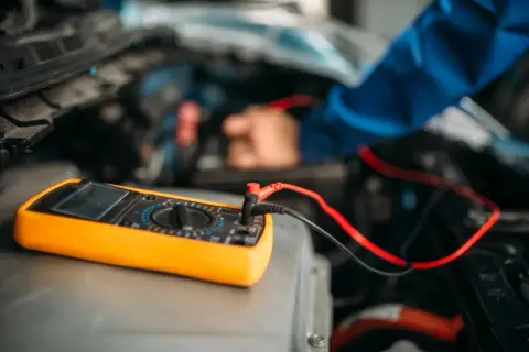 Ein Mechaniker testet die auto batterie mit einem Multimeter. Das gelbe Gerät liegt auf dem Motorraum.
