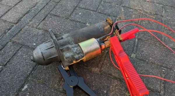 Ein defekter Anlasser, der nur klackt, liegt auf Pflastersteinen. Rote Kabel mit Klemmen sind angeschlossen.