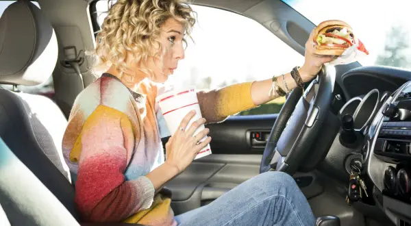 Frau trinkt und hält einen Burger beim Autofahren. Ob man beim Autofahren essen darf, ist eine Frage der Konzentration.