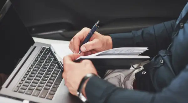 Mann schreibt im Notizbuch, während er im Auto sitzt. Er dokumentiert seinen Arbeitsweg, ob privat oder dienstlich, und nutzt dabei seinen Laptop.