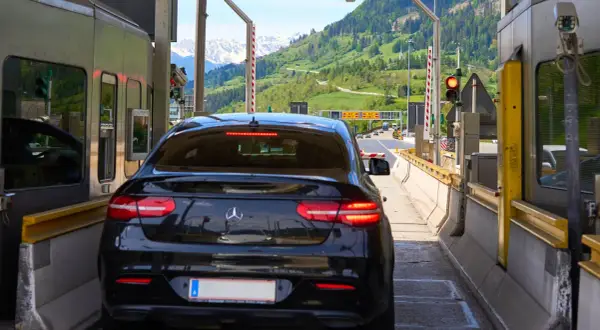 Schwarzer Firmenwagen an einer Mautstation, bereit für die Fahrt ins Ausland. Berge im Hintergrund.