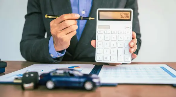 Mann mit Stift und Taschenrechner, der 9000 anzeigt. Auf dem Tisch liegen ein Auto-Schlüssel und ein Modellauto, was auf die **Erstattung Minderkilometer Leasing** hindeutet.