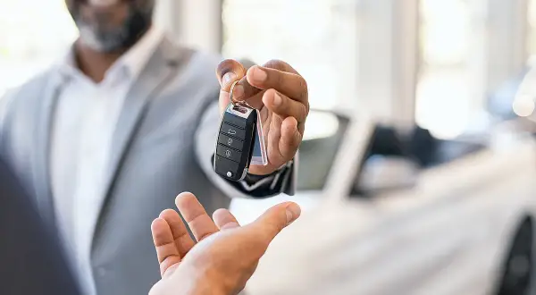 Ein Mann übergibt lächelnd Autoschlüssel. Ein neuer Wagen steht bereit, um die **Regelung umgehen** zu können und sofort loszufahren.