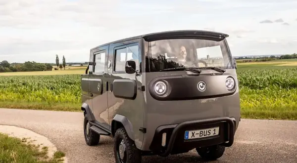 Grauer X-Bus 1 fährt auf Feldweg, Fahrer lächelt. Dieses Elektrofahrzeug ist ein Beispiel für moderne elektrische Verbraucher.