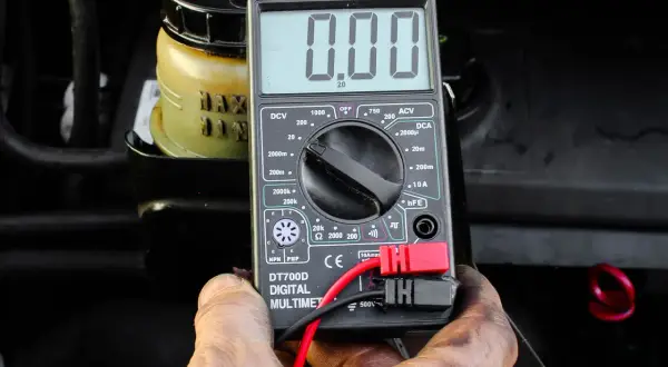 Auto springt nicht an, aber Radio geht. Ein Multimeter zeigt 0.00 an, während im Hintergrund ein Behälter mit Flüssigkeit zu sehen ist.