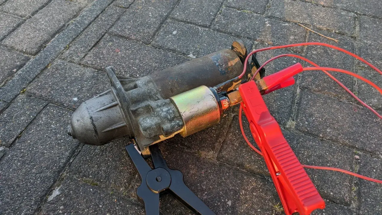 Ein defekter Anlasser, der nur klackt, liegt auf Pflastersteinen. Rote Kabel mit Klemmen sind angeschlossen.