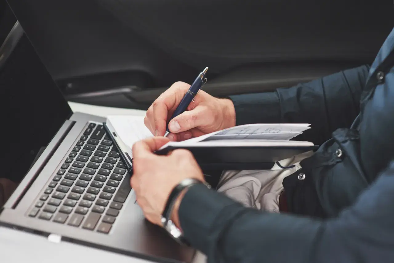 Mann schreibt im Notizbuch, während er im Auto sitzt. Er dokumentiert seinen Arbeitsweg, ob privat oder dienstlich, und nutzt dabei seinen Laptop.