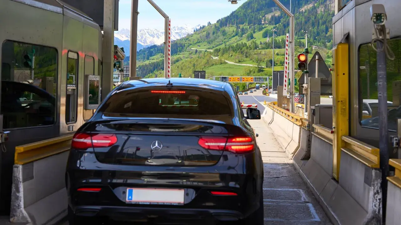 Schwarzer Firmenwagen an einer Mautstation, bereit für die Fahrt ins Ausland. Berge im Hintergrund.