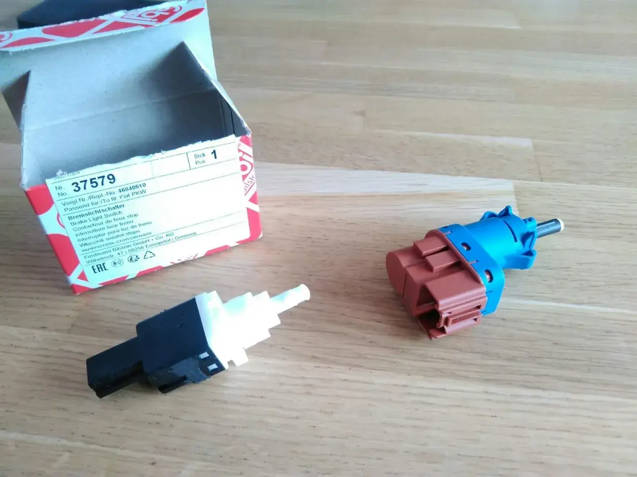 Zwei neue Bremslichtschalter liegen neben einer geöffneten Verpackung. Ein Teil ist blau und braun, das andere schwarz und weiß.