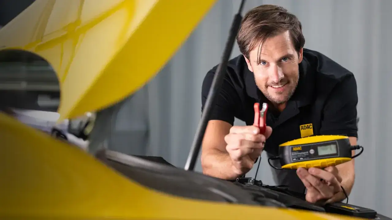 Mann mit ADAC-Kleidung h&auml;lt ein Ladeger&auml;t und eine Klemme, um die Autobatterie zu laden.