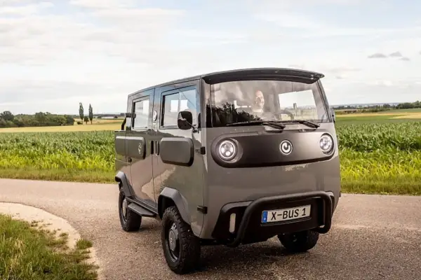 Grauer X-Bus 1 fährt auf Feldweg, Fahrer lächelt. Dieses Elektrofahrzeug ist ein Beispiel für moderne elektrische Verbraucher.