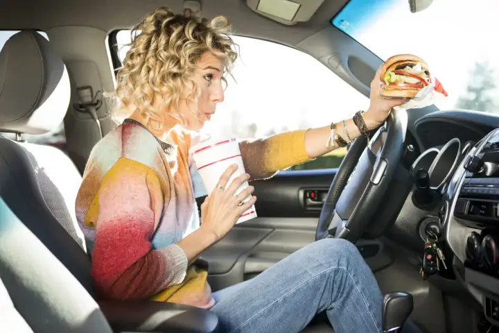 Frau trinkt und hält einen Burger beim Autofahren. Ob man beim Autofahren essen darf, ist eine Frage der Konzentration.